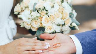 casal com mãos estendidas com as alianças na palma da mão do noivo, ao fundo, a noiva segura o buquê