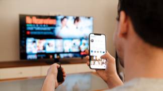 homem com celular na mão mexendo na tv com o controle remoto