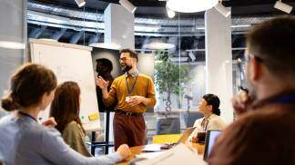 Um homem em pé explicando algo no quadro e outras pessoas sentadas namesa participando da reunião