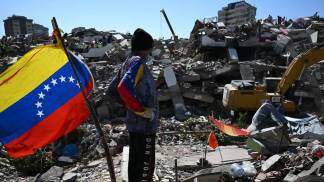 Homem encara para entulho, onde está fincada uma bandeira da Venezuela  