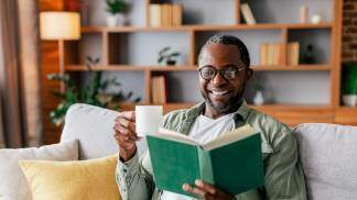 homem negro sorrindo lendo um livro no sofá e tomando café em casa