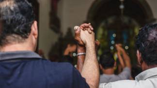 dois homens de raças diferentes vistos de costas e de mãos dadas em igreja